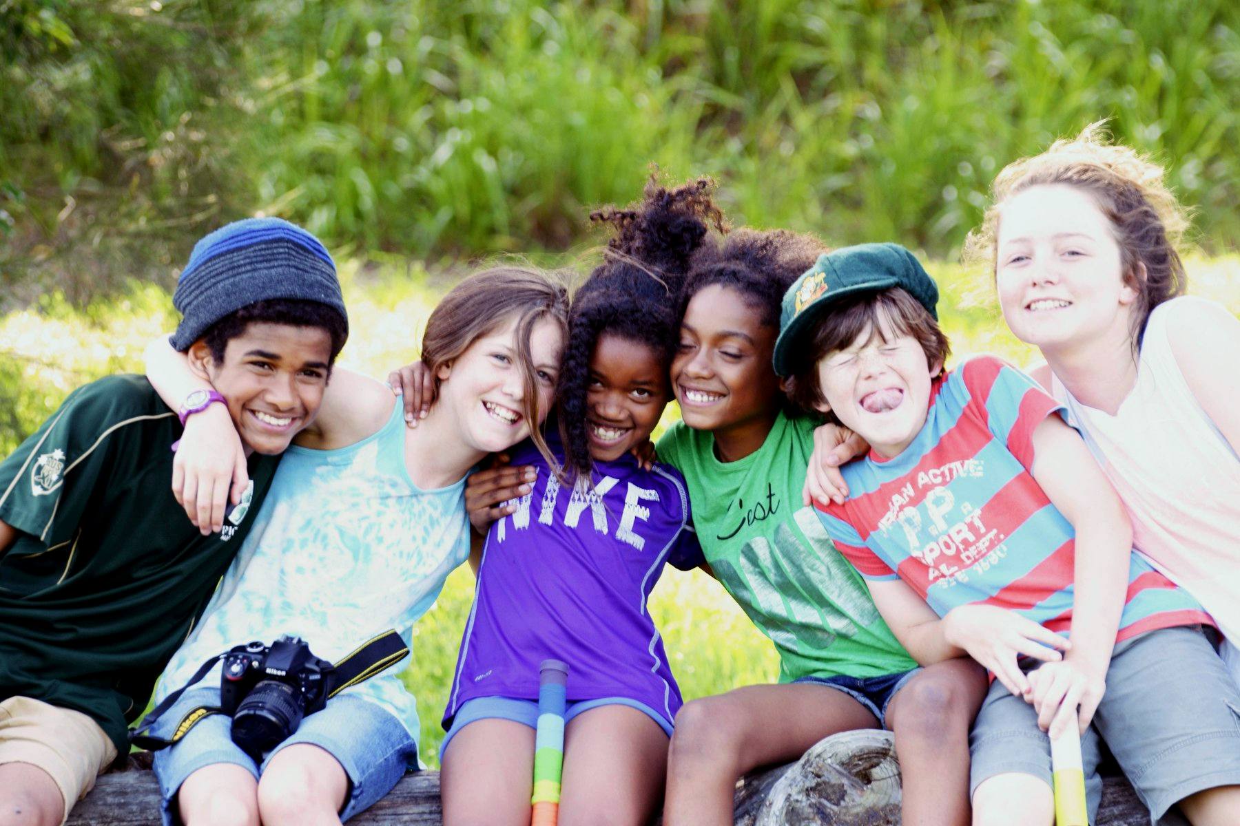 Six young friends of different backgrounds sitting on a log outdoors, arms around each other, laughing